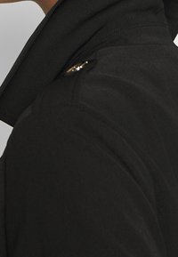 Veste noire avec un col montant, un tissu lisse, et un bouton doré brillant sur l'épaule, mettant en avant un design élégant.