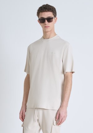 Camiseta beige de manga corta con cuello redondo y sutil logo de la marca en la esquina superior izquierda. Tejido suave con una textura lisa.