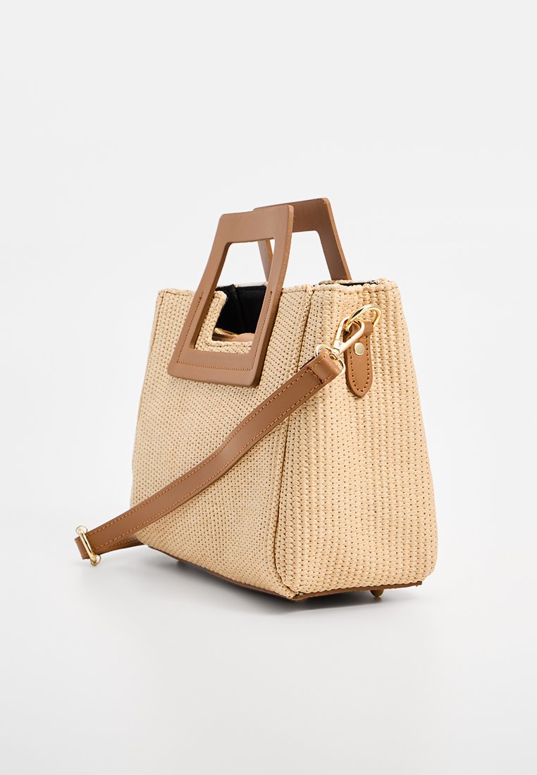Beige geweven handtas met rechthoekige bruine handvatten en een verstelbare leren schouderriem in het bruin, tegen een effen witte achtergrond.