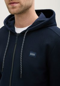 Hanorac cu fermoar navy, dintr-un material texturat, cu șnururi contrastante și un mic logo brodat pe piept. Glugă ajustabilă.