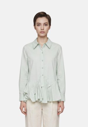 Giovane donna con capelli corti castani indossa una camicia verde chiaro a maniche lunghe con bottoni e orlo arricciato e pantaloni chiari.