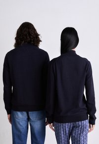 Tamno plavi pleteni pulover s rebrastim ovratnikom i manžetama, koji ima opušteni kroj. Nosi ga dvojica figura prikazana odostraga, u kombinaciji s trapericama i bermudama s uzorkom.