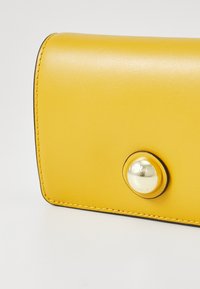 Portefeuille en cuir jaune avec une texture lisse, une forme arrondie et un fermoir à bouton pression doré bien en vue. Détails de contour noir visibles.