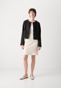 Svart cropped jakke med store knapper og to frontlommer, kombinert med et teksturert hvitt skjørt og beige leiligheter.