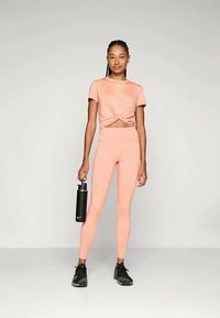 Kobieta w brzoskwiniowym stroju do ćwiczeń trzyma czarną butelkę na wodę Nike, stojąc na jasnym, jednolitym tle.