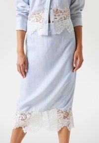 Fustă din denim albastru deschis cu dantelă albă la tiv. Prezintă o silueta ajustată și un luciu subtil. Textură netedă, până la genunchi.