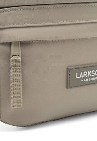 Borsa in tessuto beige con superficie testurizzata, dotata di una cerniera e una patch rettangolare con logo. Il design è minimalista con linee pulite.