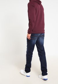 Donkerrode met capuchon trui, blauwe denim jeans, witte sneakers met blauwe accenten. De jeans hebben achterzakken en een rechte pijp.