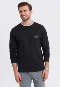 Lächelnder Mann in einem schwarzen Langarmshirt mit kleinem "EX"-Logo auf der Brust und grauen Nadelstreifenhosen, stehend vor einem weißen Hintergrund.