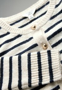 Flødefarvet sweater med marineblå striber, blød tekstur, ribbede ærmekanter og to gyldne knapper ved halsudskæringen. Mærket angiver "THE SET," størrelse medium.