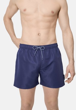 Hombre sin camiseta que lleva pantalones cortos de baño azul marino con cordón frontal, de pie contra un fondo claro y liso.