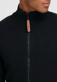 Suéter negro de punto con cremallera y alto cuello, con un tirador de cremallera de cuero en tono marrón claro y textura acanalada. Vista en primer plano.