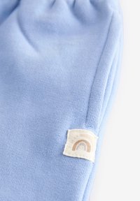 Hellblauer Strickstoff mit strukturierter Oberfläche. Am Saum befindet sich ein eingenähtes Etikett mit einem Regenbogendesign in neutralen Farbtönen.