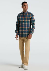 Man staat in beige broek, witte sneakers en een blauw, oranje en groene geblokte blouse, kijkend naar rechts.