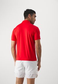 Red polo shirt made of lightweight fabric with a collared design, short sleeves, and a smooth texture, worn with white shorts.
