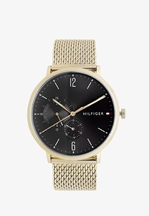 Auksinė tinklinė dirželio laikrodis su juodu ciferblatu, turi subciferblatą, skaitmeninius valandų žymeklius ir plonas auksines rodykles. Logotipas aiškiai matomas.