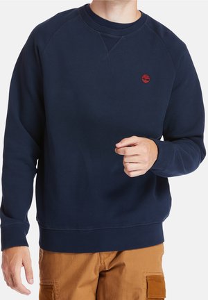 Homme portant un sweat-shirt bleu marine à col rond avec un petit logo d'arbre rouge sur la poitrine gauche et un pantalon marron clair, debout devant un fond blanc.