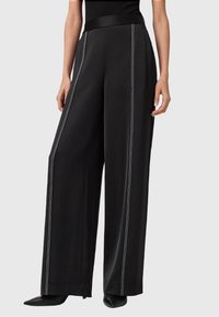 Zwarte wijde broek met witte verticale stiksels, gedragen met zwarte puntige hakken en een zwart mouwloos topje tegen een effen achtergrond.