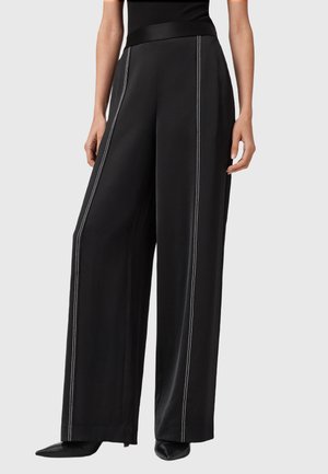Pantaloni neri a gamba larga con cuciture verticali bianche indossati con tacchi a punta neri e un top senza maniche nero su uno sfondo neutro.