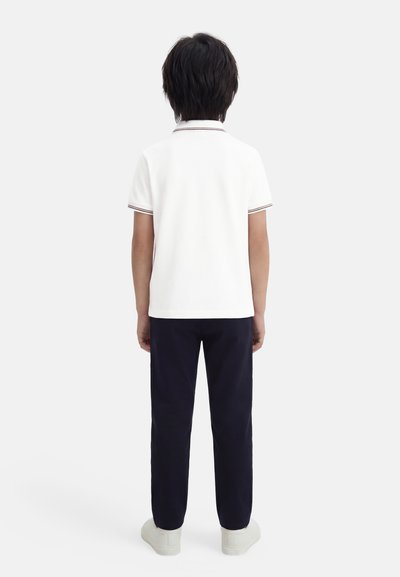 Enfant aux cheveux foncés, vu de dos, portant un polo blanc à manches courtes, un pantalon foncé et des baskets blanches, sur un fond uni.