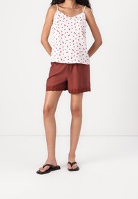 Débardeur blanc avec motif de cerises rouges, bretelles fines, associé à un short couleur rouille avec un ourlet en dentelle festonné. Tongs noires.