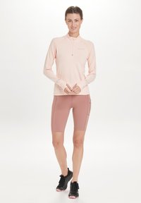 Hellrosa langärmliges Zip-Top mit Daumenlöchern, kombiniert mit rosa Fahrradhosen mit seitlicher Tasche. Schwarze Sportschuhe.