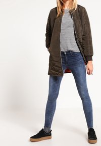 Chaqueta de plumas larga color verde oliva con textura suave, cuello redondo, camiseta de rayas gris debajo, jeans skinny azul y zapatillas negras de plataforma.