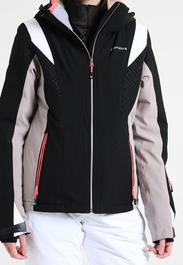 Femme portant une veste de ski noire, beige et blanche avec des détails de fermeture éclair roses et un pantalon de neige blanc.