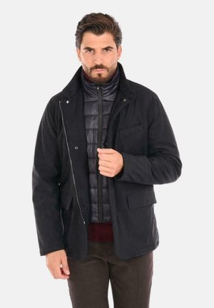 Giacca esterna nera sopra un gilet imbottito scuro, con chiusura a zip e bottoni, tasche anteriori e tessuto testurizzato. Vestibilità casual con colletto.