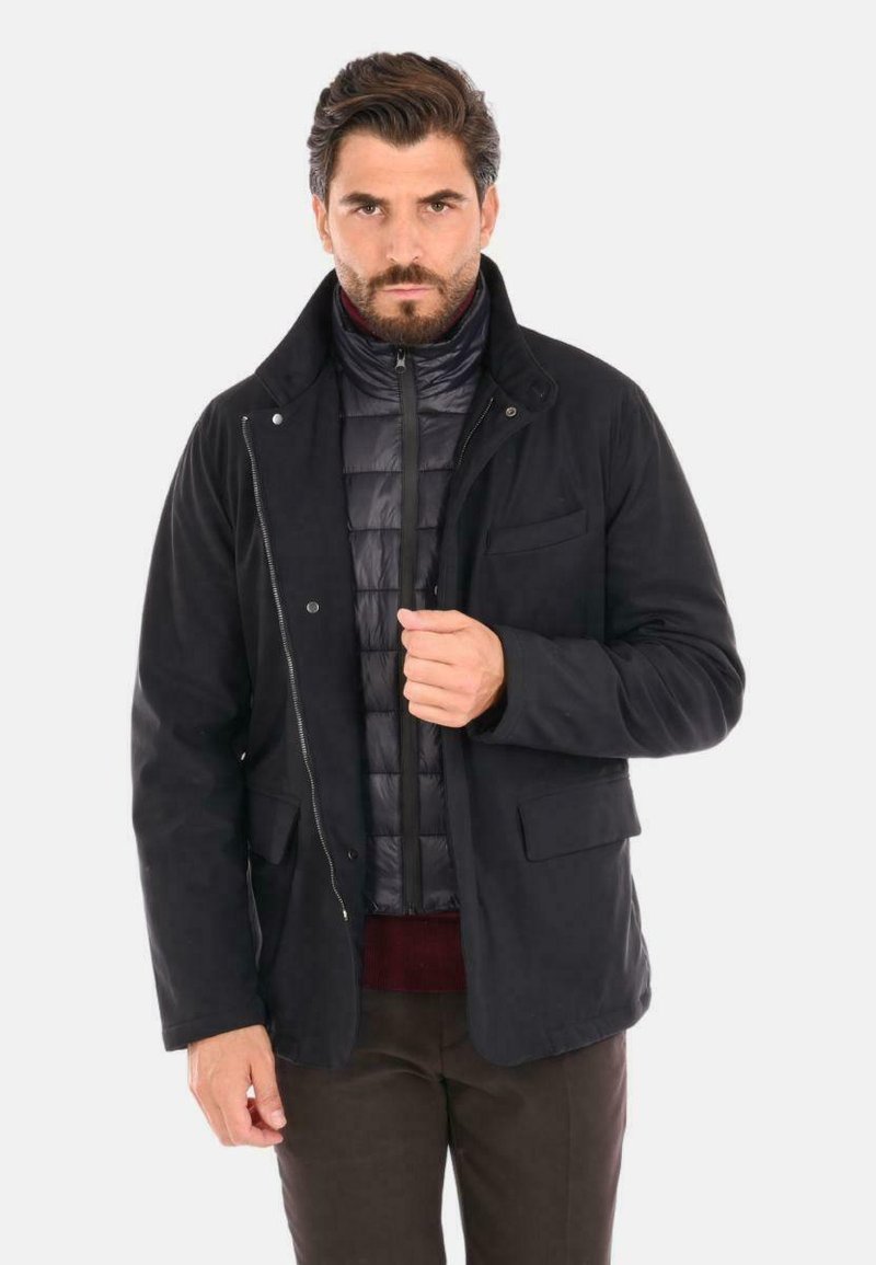 Giacca esterna nera sopra un gilet imbottito scuro, con chiusura a zip e bottoni, tasche anteriori e tessuto testurizzato. Vestibilità casual con colletto.