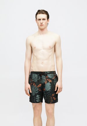 Jeune homme debout, torse nu, portant un short de bain noir avec des motifs de feuilles tropicales vertes et orange, sur un fond clair uni.