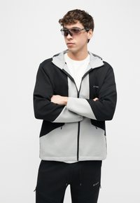 Νεαρός άνδρας που φοράει μαύρη και γκρι ζακέτα με φερμουάρ και μαύρο παντελόνι, με τα χέρια σταυρωμένα, κοιτάζοντας πλάγια, φορώντας σκουρόχρωμα γυαλιά ηλίου τύπου wraparound.