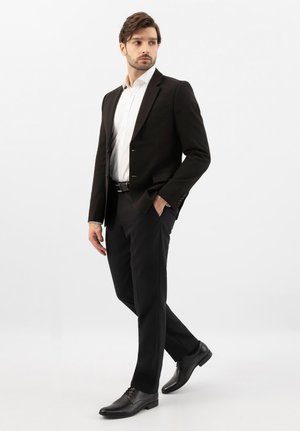 Mann im schwarzen Anzug, weißem Hemd und schwarzen Lederschuhen steht mit einer Hand in der Tasche und schaut seitlich auf weißem Hintergrund.