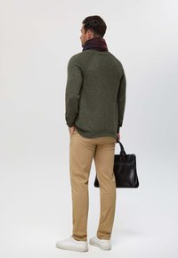 Pull en tricot vert foncé, pantalon beige et baskets blanches. La mannequin porte un sac en cuir noir. L'écharpe présente des couleurs rouge foncé et bleu marine.