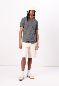 Black and white striped t-shirt, relaxed fit, paired with cream-colored shorts. Complemented by a beige bucket hat and white socks with sneakers.