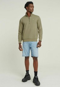 Felpa verde oliva con zip e colletto rotondo, abbinata a pantaloni shorts in denim azzurro e sneakers nere, in piedi davanti a uno sfondo neutro.