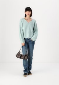 Pull en maille texturée bleu clair avec décolleté en V, associé à un jean bleu. Le mannequin tient un petit sac à main marron foncé avec des compartiments.