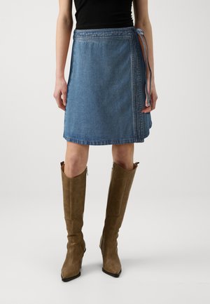 Femme portant une jupe portefeuille en jean bleu jusqu'aux genoux et de grandes bottes marron à talons, debout devant un fond clair uni.