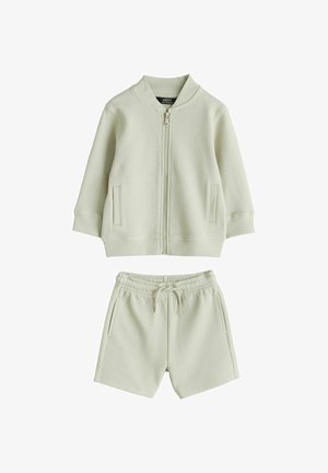 Svijetlozeleni pleteni trenirka s jaknom na patentni zatvarač i odgovarajućim kratkim hlačama. Jakna ima dva bočna džepa i rebraste manžete. Tkanina je teksturirana.