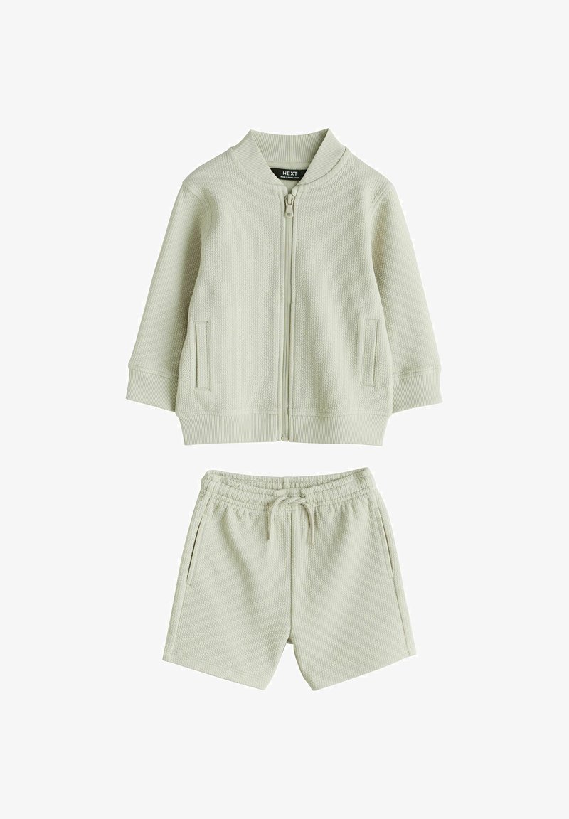 Ensemble de survêtement en maille vert clair comprenant une veste zippée et un short assorti. La veste est dotée de deux poches latérales et de poignets côtelés. Tissu texturé.