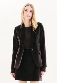 Blazer nero su misura con una texture liscia, chiusura a un bottone e rever a scollatura, abbinato a una gonna nera coordinata.