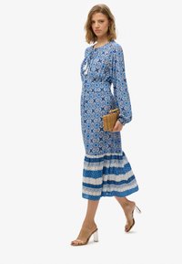 Niebieska sukienka maxi w wzory z bufiastymi rękawami, ściągnięta w pasie oraz z rozkloszowanym dołem. Nosi ją w połączeniu z przezroczystymi sandałami na obcasie oraz plecioną torebką.