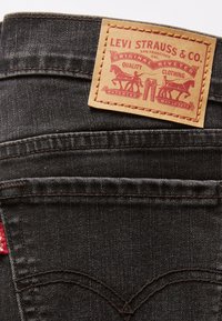 Jeans de mezclilla negra con un parche de cuero marrón que lleva una marca roja, costuras doblemente cosidas y una pequeña etiqueta roja en el bolsillo trasero.