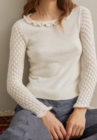 Kremowy sweter z falbankowym dekoltem, długimi koronkowymi rękawami i prążkowanym dołem. Połączony z szarymi spodniami, ukazujący miękką teksturę i skomplikowane wzory.