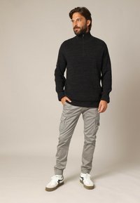 Man met een baard in een zwart gebreid pullover met een halve rits, lichtgrijze cargo-broek en witte sneakers, staand met zijn handen in zijn zakken.
