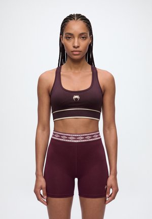 Une femme porte un soutien-gorge de sport bordeaux et un short ajusté assorti sur un fond uni clair.