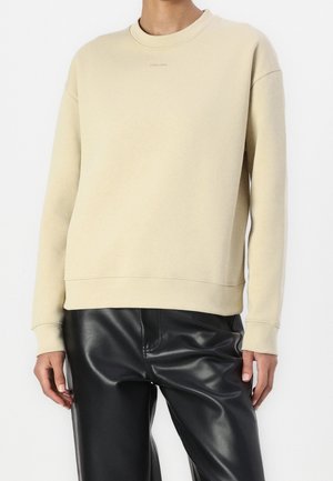 Felpa beige con colletto e polsini a coste, caratterizzata da un sottile logo Calvin Klein. Abbinata a pantaloni in ecopelle neri per creare contrasto.