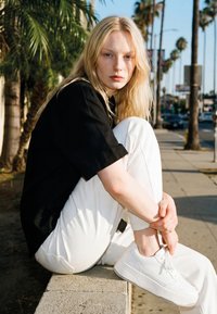 Junge Frau mit langem blondem Haar, die ein schwarzes Hemd und weiße Hose trägt, sitzt auf einer Betonmauer an einem sonnigen Bürgersteig mit Palmen.