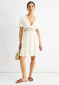 Robe blanche à manches courtes, décolleté en V profond, accents en dentelle, ourlet en couches, assortie avec des sandales marron et un sac en osier. Tissu lisse et léger.