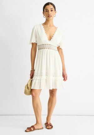 White dress with short sleeves, deep v-neck, lace accents, tiered hem, paired with brown sandals and a woven bag. Smooth, lightweight fabric.